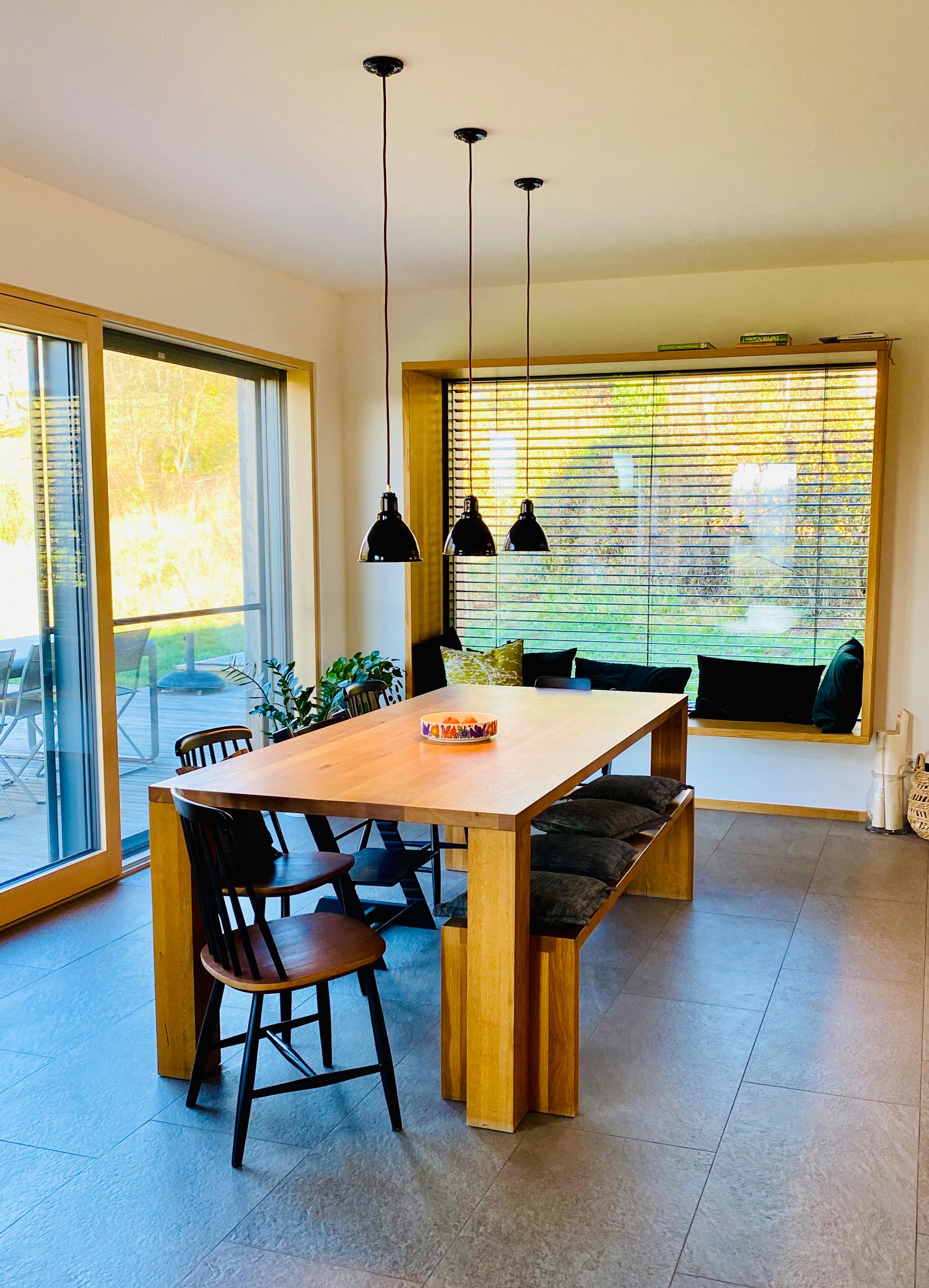 Esstisch Beleuchtung schwarz Ein moderner Wohnraum mit großen, bodentiefen Fenstern. In der Mitte steht ein großer Esstisch aus hellem Holz. Über dem Tisch hängen drei kleine, schwarze Lampen aus Emaille