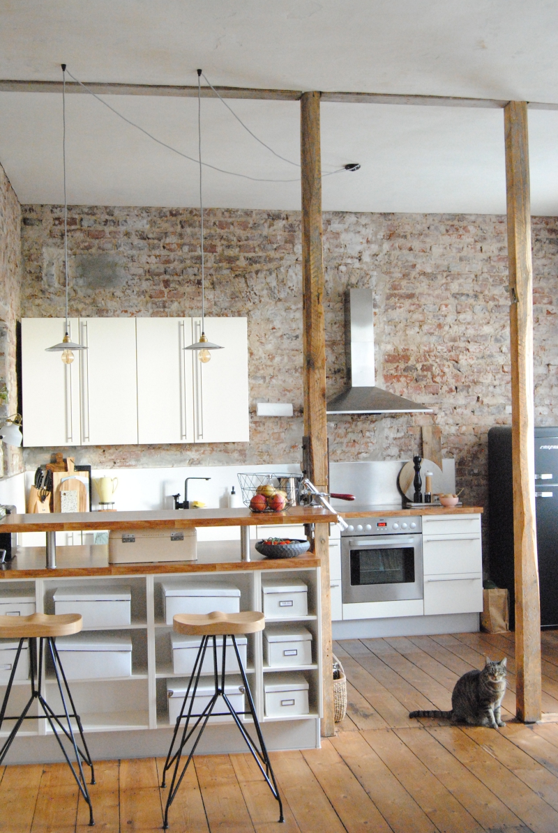 Küche im Industriestil Küchenzeile in einer Altbauwohnung mit einer freigelegten Wand aus Backsteinen. Die Küchenfronten sind weiß und es hängen zwei kleine Fabriklampen über einer Theke aus Holz