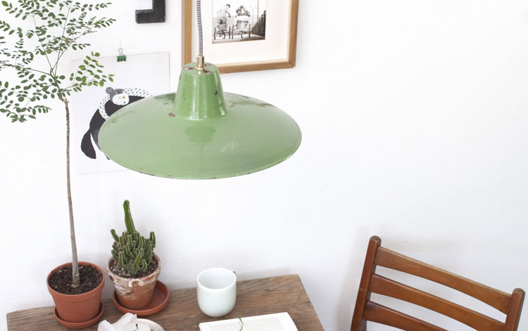 grüne Emaillelampe über Esstisch Im Bildausschnitt ist ein Teil eines altes Holztisches zu sehen, darauf stehen zwei Grünpflanzen und eine Kaffeetasse. Über dem Tisch hängt eine Lampe aus grünem Emaille