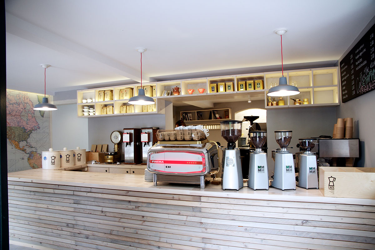 Kaffeetheke in einem Café in Korfu Eine Theke aus hellem Holz. darauf stehen eine Siebträger-Kaffeemaschine sowie mehrere Kaffeemühlen. Über der Theke hängen drei graue Fabriklampen an roten Kabeln