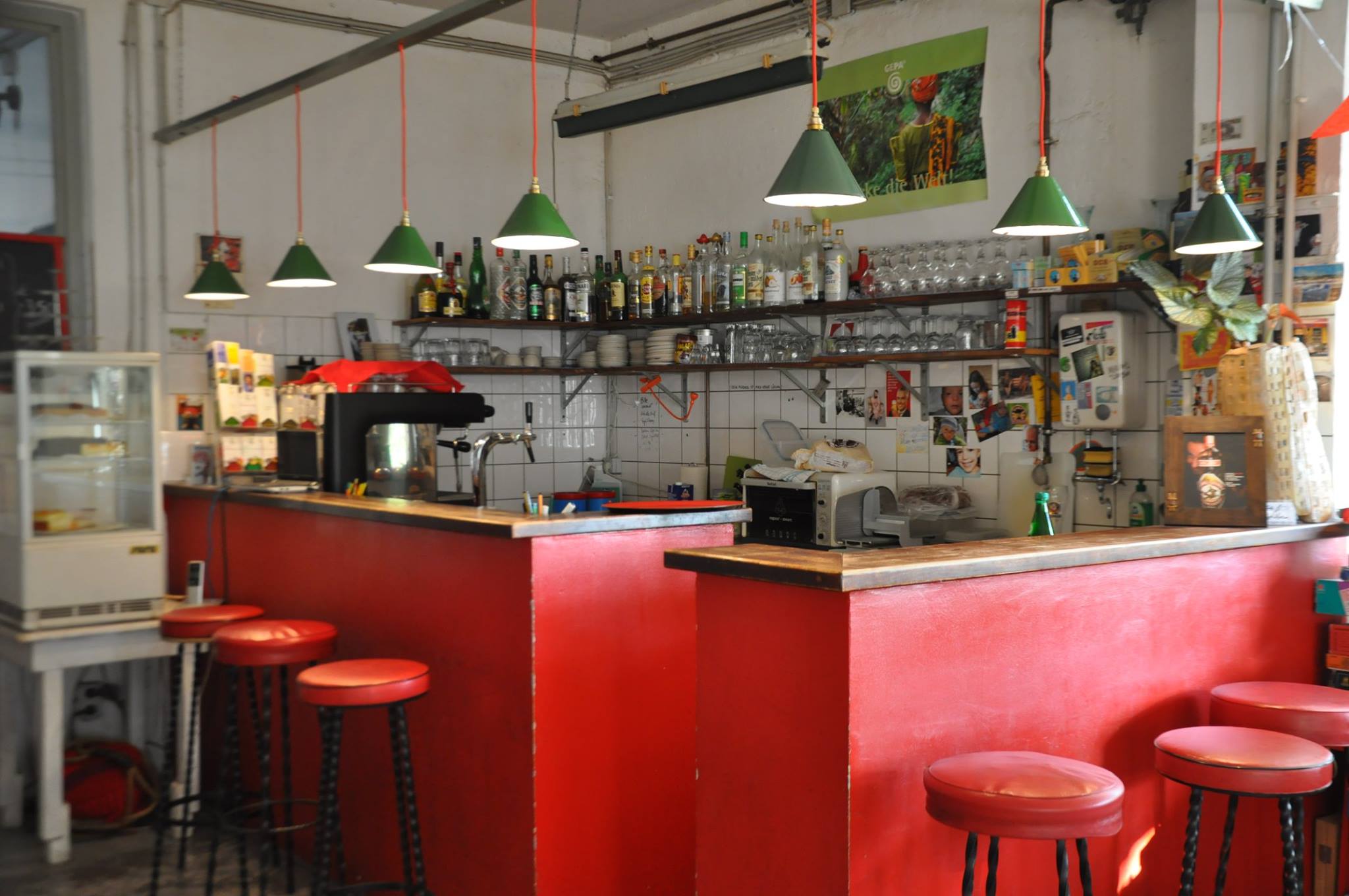 Rote Bar im Café Libre eine rote Theke in einer Bar. Darüber hängen 7 kleine kegelförmige Lampen aus grünem Emaille. Vor der Theke stehen Barhocker, die ebenfalls rot sind. Hinter der Theke befinden sich Regale an den Wänden, die mit verschiedenen Flaschen gefüllt sind.
