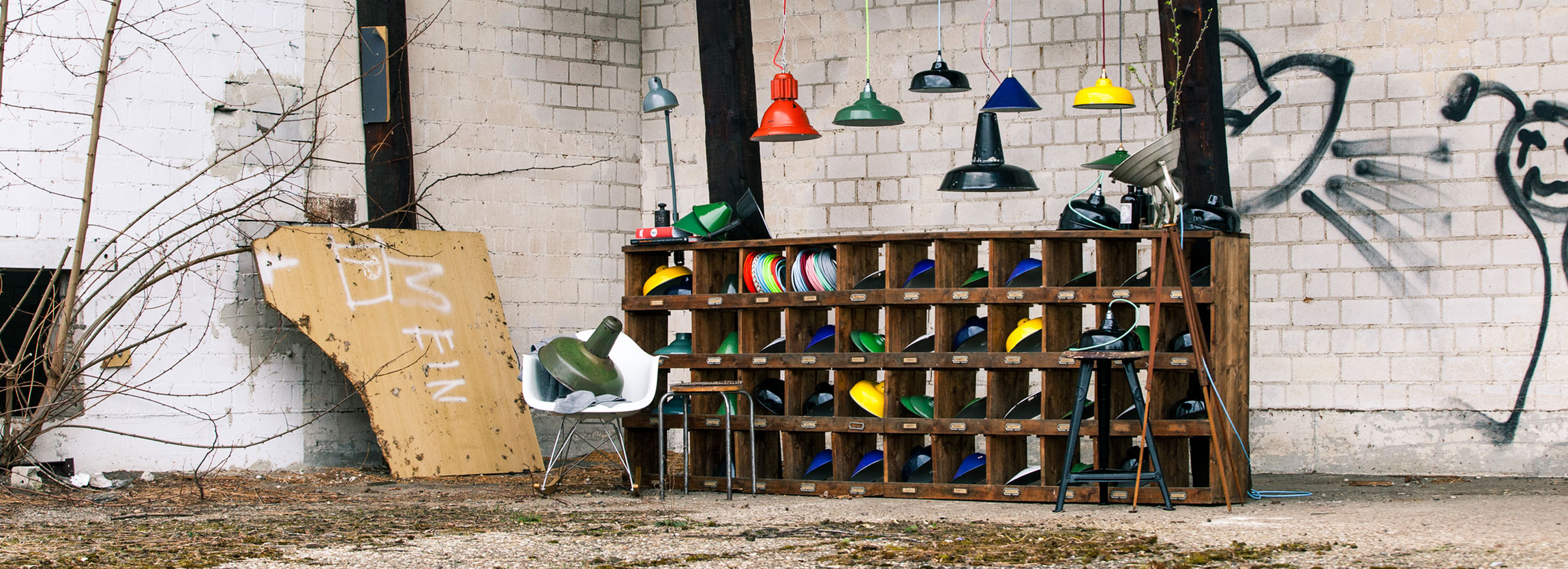 fullscreen_start Ein verlassenes Fabrikgelände. Vor einer weißen Steinwand, die mit schwarzen Graffitis beschmiert ist, steht ein großes Holzregal mit vielen Fächern. In den Fächern sind bunte Lampenschirme und bunte Stoffkabel. Über dem Regal hängen alte Fabriklampen in