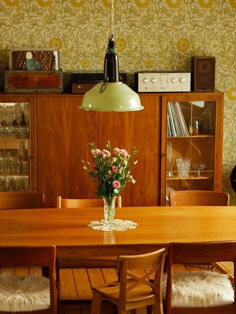 Esszimmer von den Fünfzigerjahren inspiriert Blick in ein Esszimmer. An einer Wand mit grüner, geblümter Tapete, steht ein Highboard-Schrank aus den 50er Jahren. Davor ein Holztisch und zwei Holzstühle - ebenfalls aus den Fünfzigern. Auf dem Tisch steht eine Glasvase mit Blumen. Über dem Tisch hängt