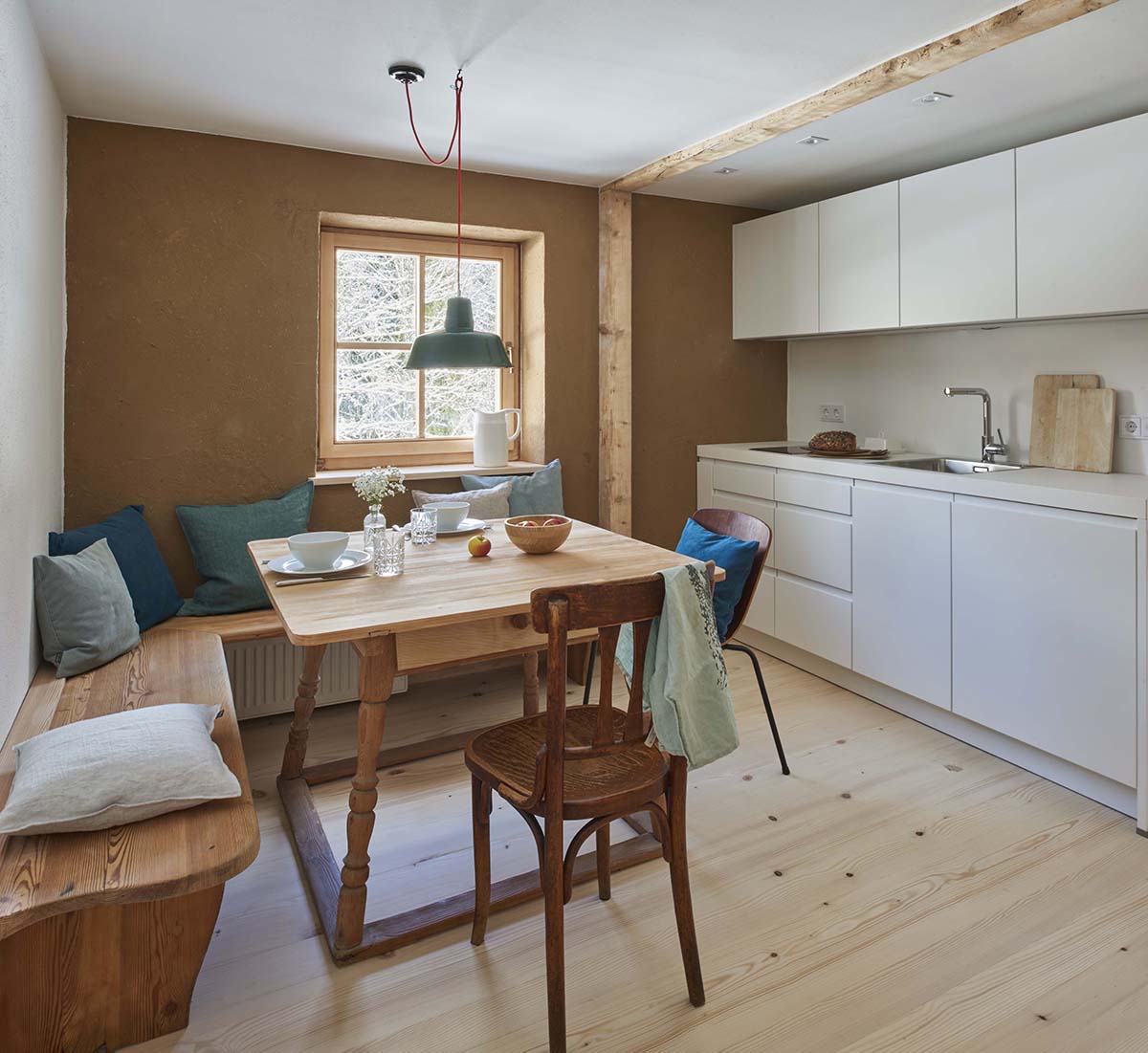 Moderne Wohnküche mit Bauernmöbeln Blick in eine Wohnküche mit kleiner, weißer Küchenzeile, hellbraunen Wänden und einem Holztisch mit Eckbank und Holzstühlen. Über dem Tisch hängt eine alte, grüne Werkstattlampe mit rotem Kabel